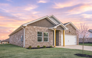 SpruceExterior2 The Spruce floor plan by Maplewood Homes featuring a single-story brick exterior, two-car garage, covered front porch, blooming spring tree, and soft sunset sky.